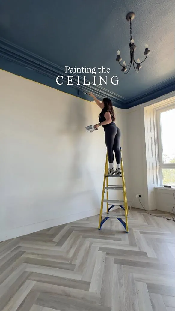 Emma on a ladder painting a ceiling deep blue above a herringbone floor