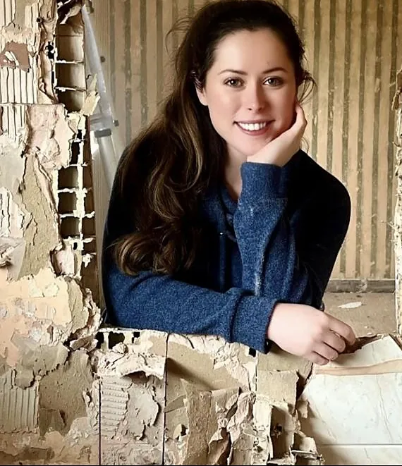 Emma smiling, leaning on a half-stripped wall of old lath and plaster