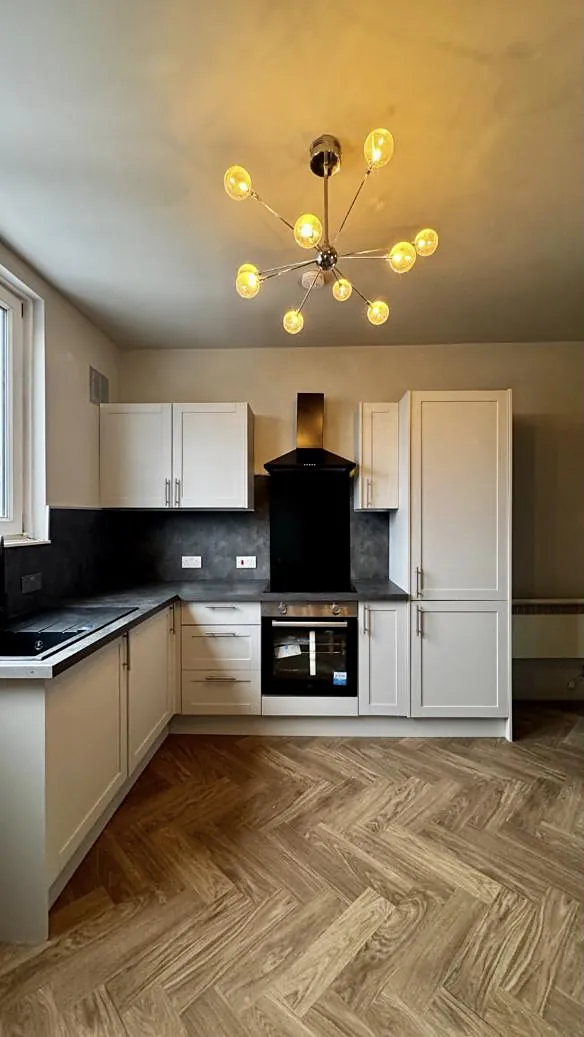 A cream shaker kitchen with a dark worktop, a glass chandelier and a warm oak herringbone floor